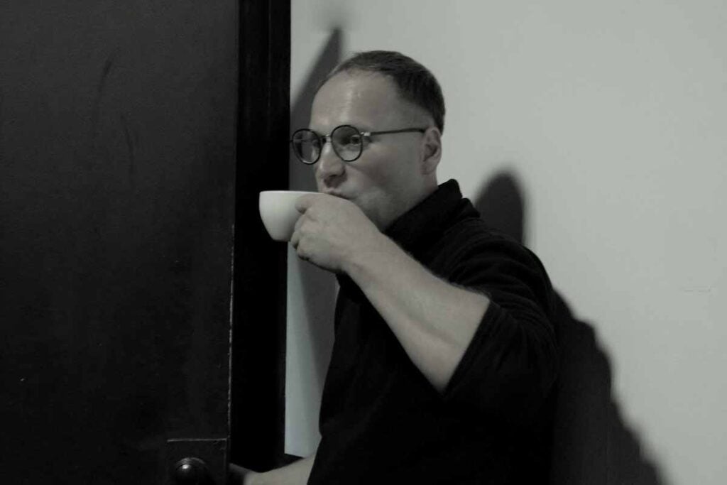 Maks Ivantsov står lutad mot en vägg inomhus och dricker vad ser ut att vara en kopp kaffe.